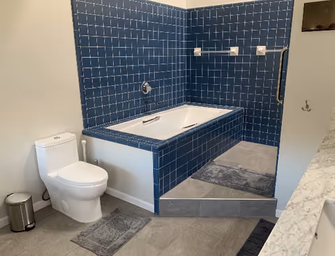 Bathroom with a blue-tiled recessed bathtub, toilet, and gray floor mats.