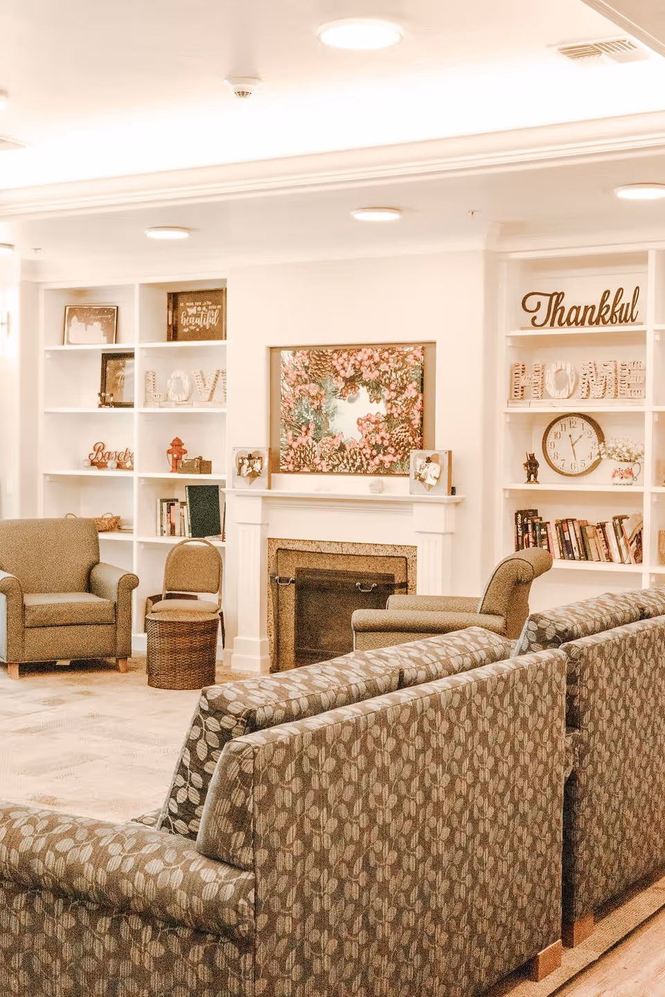 Cozy communal living room with patterned sofas and armchairs facing a fireplace flanked by built-in shelves filled with decor.
