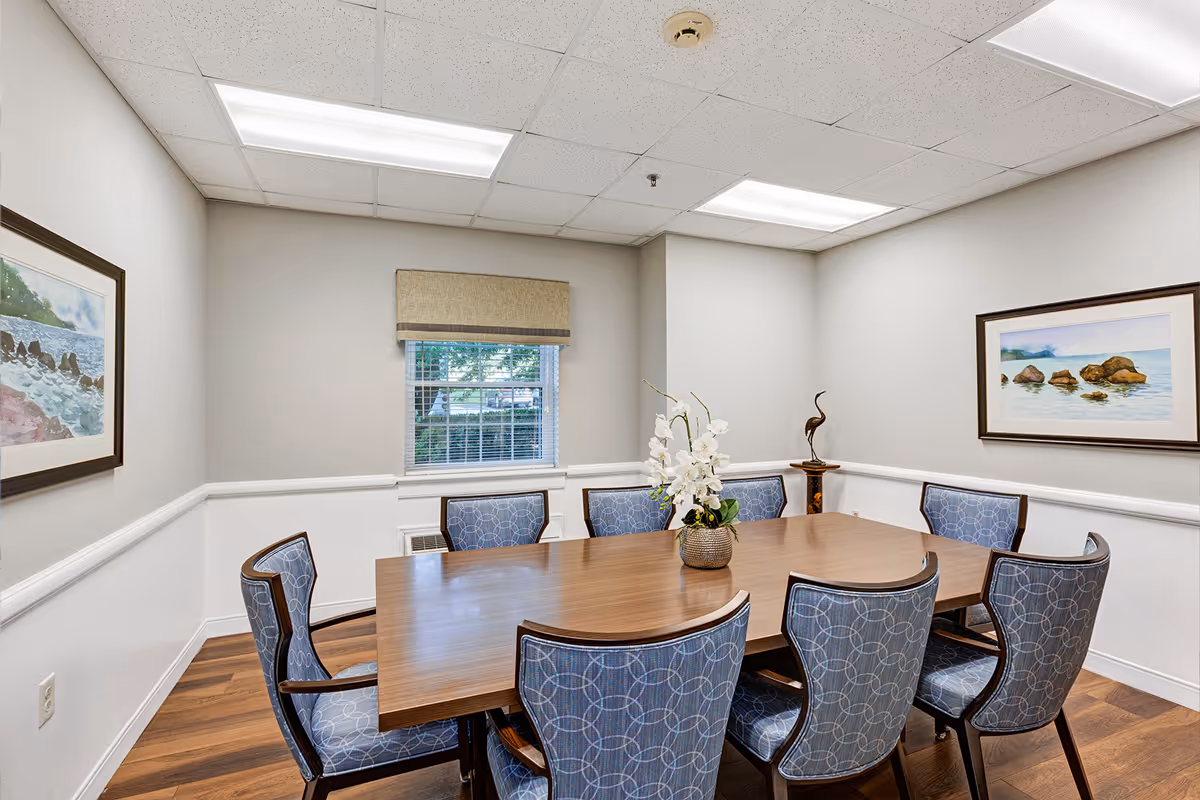 A small meeting or dining room with a rectangular wooden table surrounded by eight blue patterned chairs. The room has light gray walls with white wainscoting, two framed landscape paintings on the walls, a window with blinds and a beige valance, and a decorative vase with white flowers on the table. The floor is wood, and the ceiling has fluorescent lighting panels.