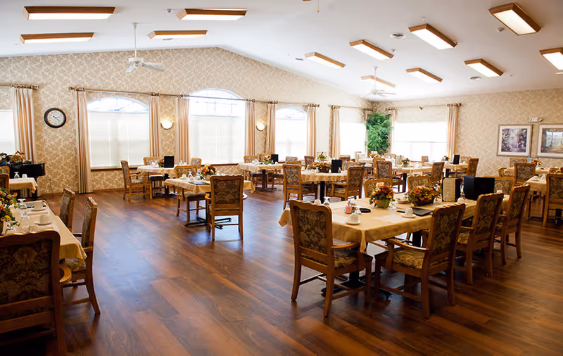 A spacious dining room with multiple tables covered with beige tablecloths, each set with cups, plates, and floral centerpieces. The room has wooden flooring, large windows with beige curtains, and decorative wall sconces. There are upholstered wooden chairs around the tables and framed artwork on the walls.