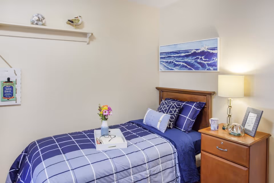 A neatly made single bed with blue and white checkered bedding and multiple pillows in a senior living facility room. A small wooden nightstand beside the bed holds a lamp, a framed quote, a cup, and a decorative glass item. Above the bed is a painting of ocean waves, and a small shelf on the adjacent wall displays decorative items including a bird figurine and seashells. A white tray with a vase of flowers and reading glasses rests on the bed.