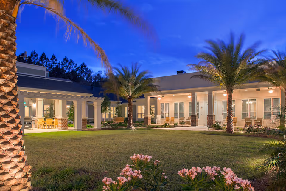 Evening view of a landscaped courtyard with palm trees, lawn, and covered patio seating at a senior living facility.