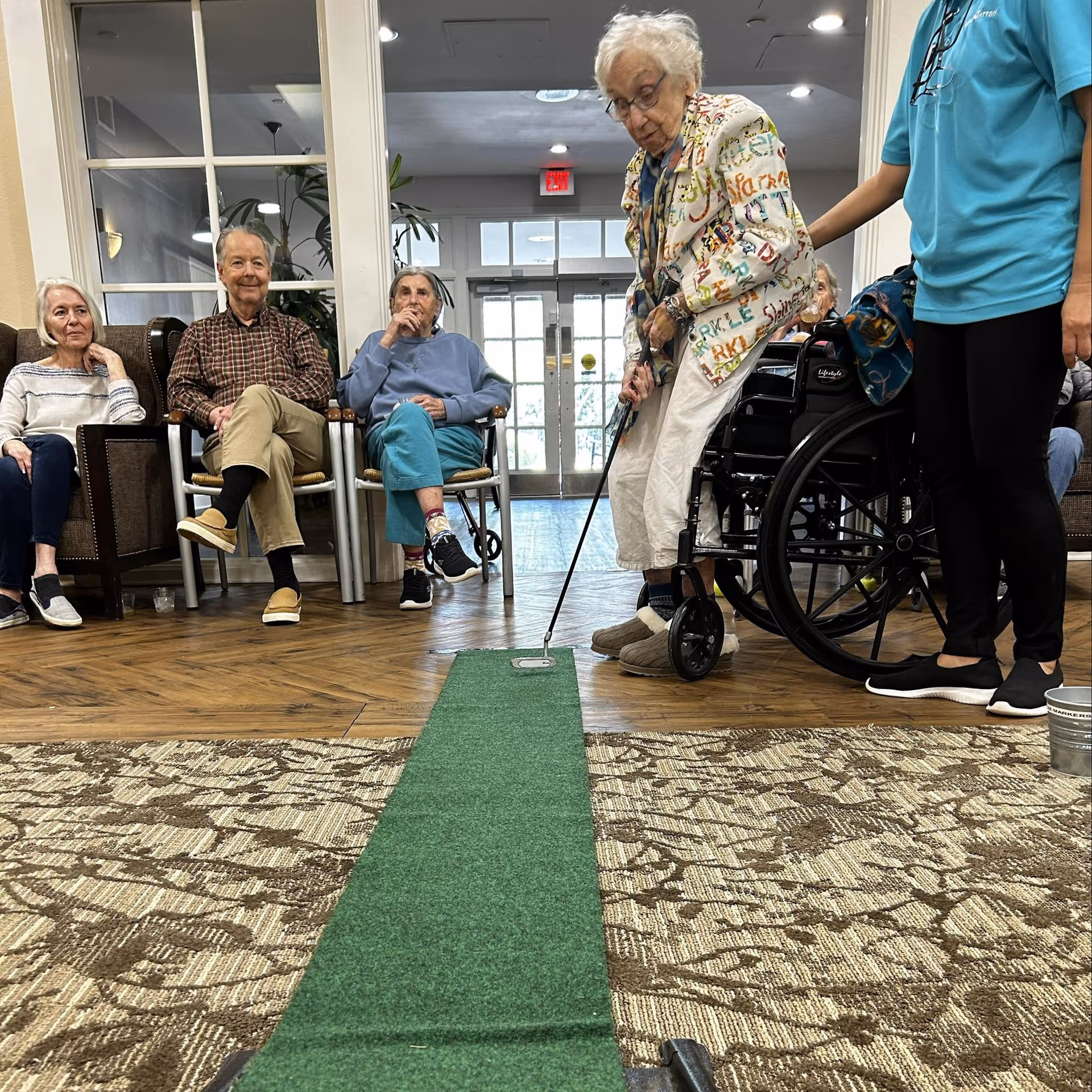 An elderly woman is playing indoor mini-golf on a green putting mat in a common area. She is holding a golf club and aiming at a golf ball near a hole. Several elderly people are seated in chairs watching her, and a caregiver stands nearby providing support. The room has a patterned carpet and large windows in the background.