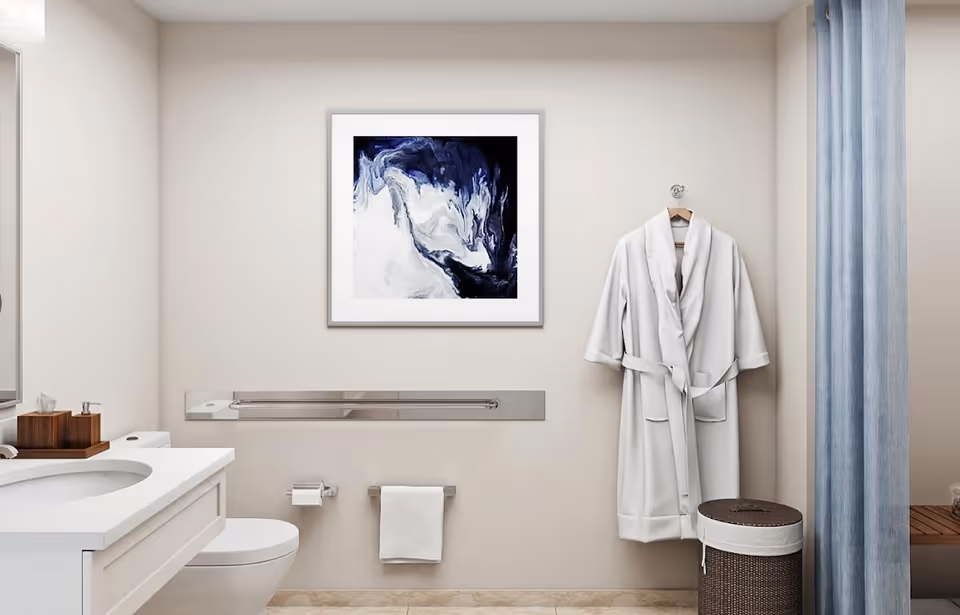 A clean and modern bathroom with a white countertop sink, a toilet, a white towel hanging on a rack, a white bathrobe hanging on a hook, a wicker laundry basket, a blue shower curtain, and a framed abstract painting with blue and white colors on the wall.