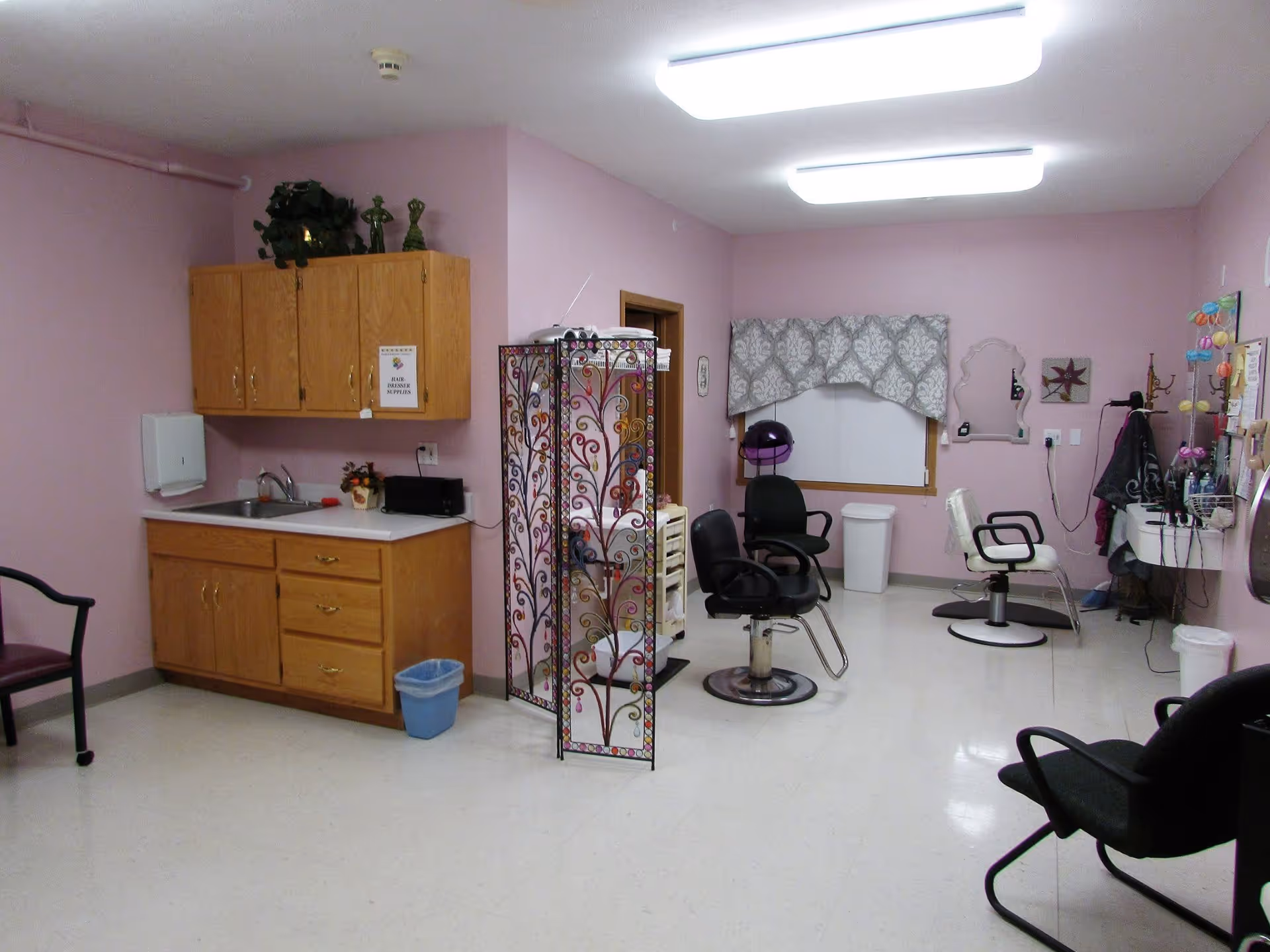 Interior of a hair salon room with pink walls, wooden cabinets, a sink, a decorative folding screen, salon chairs, a hair dryer, and various hair styling tools and accessories.