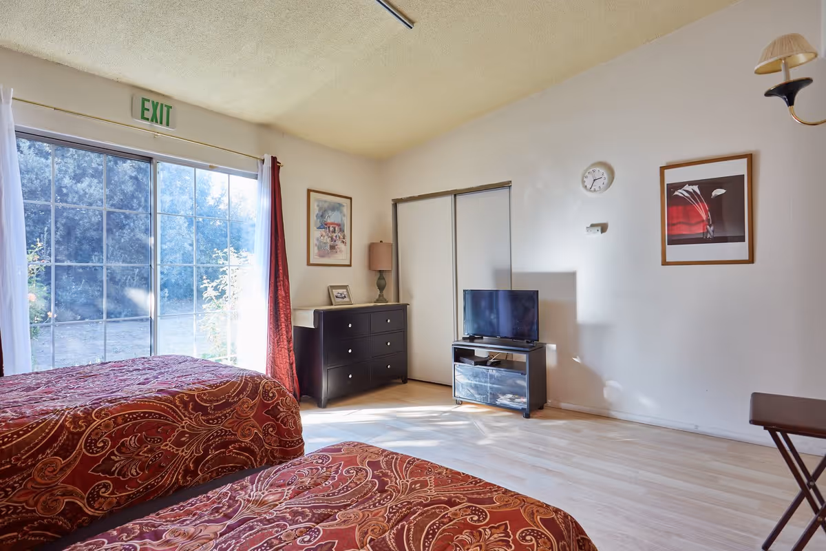 A bright bedroom with two beds covered in red patterned bedspreads. The room has a large window with white and red curtains letting in natural light. There is a black dresser with a lamp and framed pictures on top, a small TV on a stand, a wall clock, and two framed pictures on the walls. The floor is light wood, and there is an exit sign above the window.