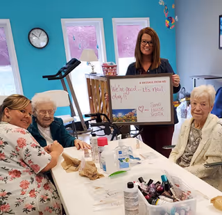 A group of three elderly women and a caregiver sitting around a table in a bright room with teal walls. The caregiver is holding a whiteboard that reads 'We're great... it's mail day! ❤️ - Sandy, Louise, Muriel.' The table has various items including nail polish bottles and hand sanitizer, suggesting a nail care activity.