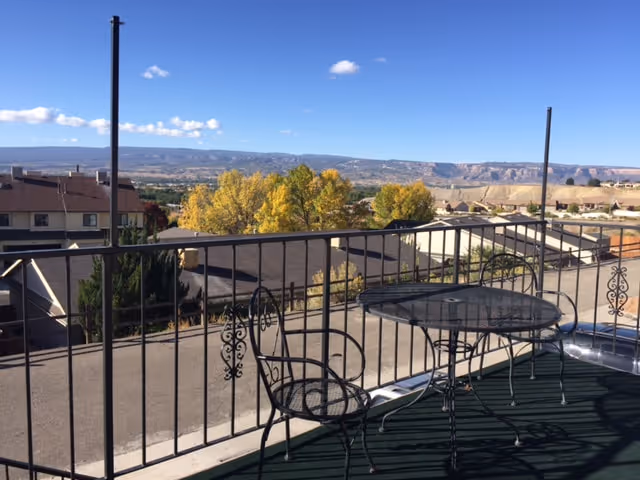 Outdoor balcony area with a black metal table and two matching chairs overlooking a residential neighborhood with trees and distant hills under a blue sky with a few clouds.