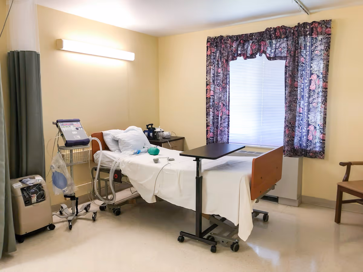 A hospital-style room with a single bed equipped with white sheets and pillows, a bedside table with medical equipment, an overbed table, floral curtains covering a window, and a wooden chair in the corner.