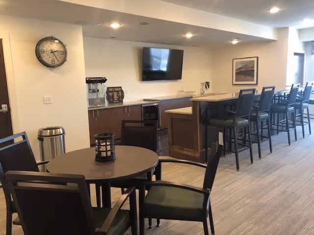 Interior view of a senior living facility common area with a round table and chairs in the foreground, a wall clock, a countertop with bar stools, a mounted flat-screen TV, and a popcorn machine on the counter.