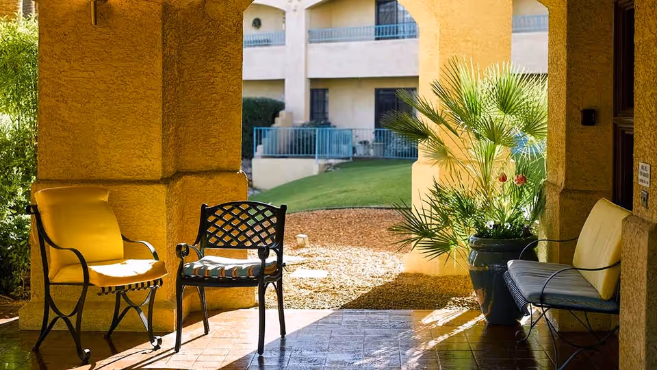 A shaded outdoor seating area with two metal chairs, one with a yellow cushion and one with a patterned cushion, and a metal bench with yellow cushions. The area is framed by yellow stucco columns and overlooks a grassy courtyard with a potted plant and a building with balconies in the background.