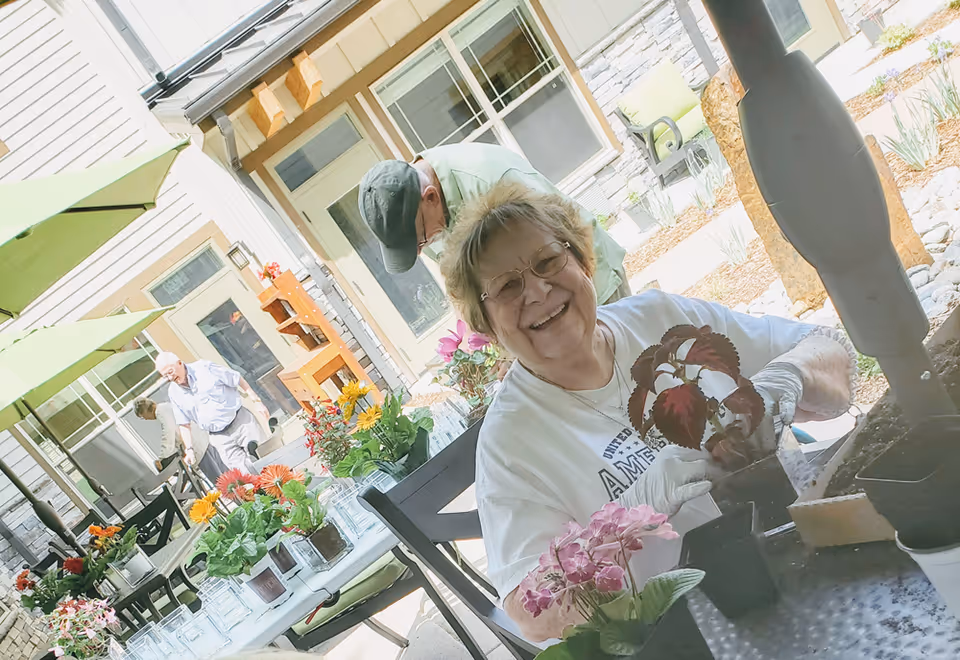An elderly woman wearing glasses and gloves is smiling while planting flowers at a table outdoors. Other elderly individuals are also engaged in gardening activities in the background, with green umbrellas providing shade. The setting appears to be a courtyard or patio area of a senior living facility.