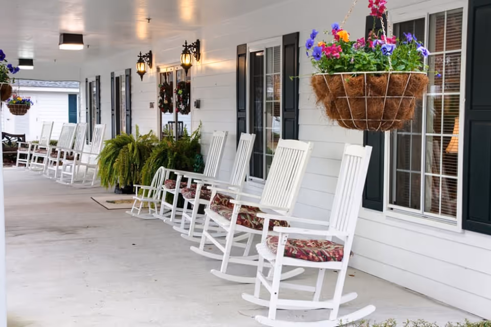 A covered porch area with a row of white rocking chairs with floral cushions, hanging flower baskets, green plants, and wall-mounted lantern-style lights. The porch is attached to a white building with black shutters and windows.