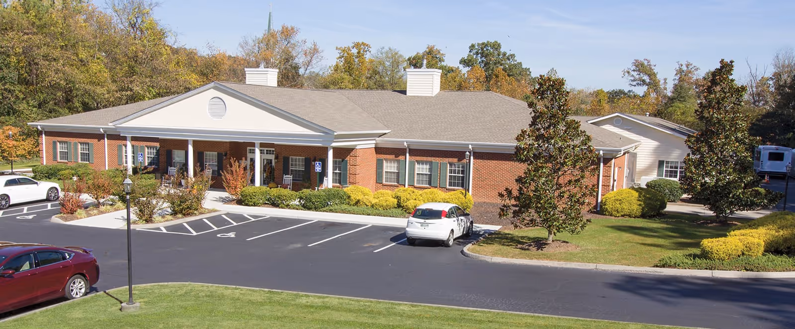 Exterior view of a single-story brick building with a covered entrance supported by white columns. The building is surrounded by a parking lot with several cars parked, landscaped bushes, trees, and a well-maintained lawn under a clear sky.