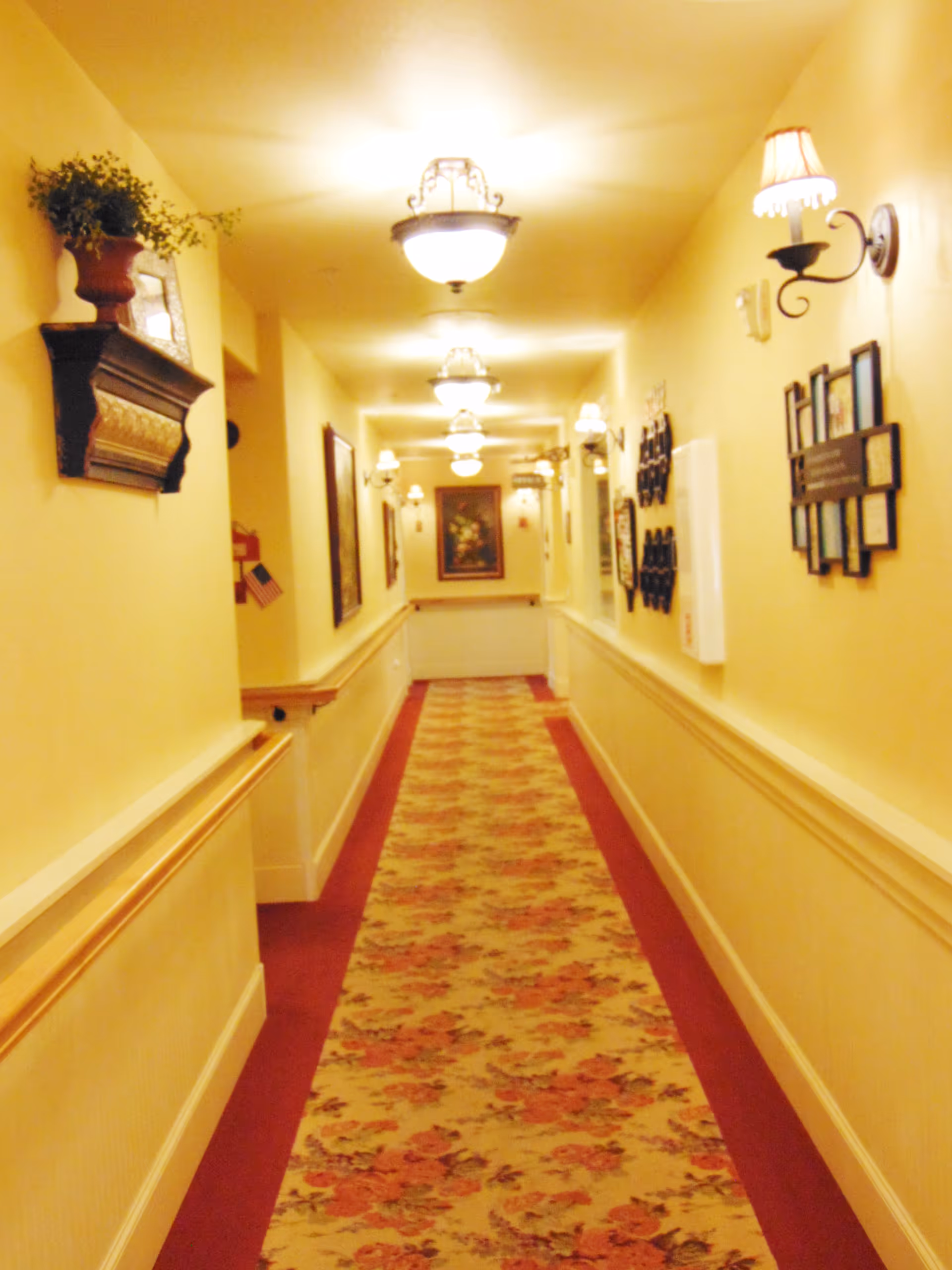 A long carpeted hallway in an assisted living facility lined with wall sconces, framed pictures, and overhead lights.