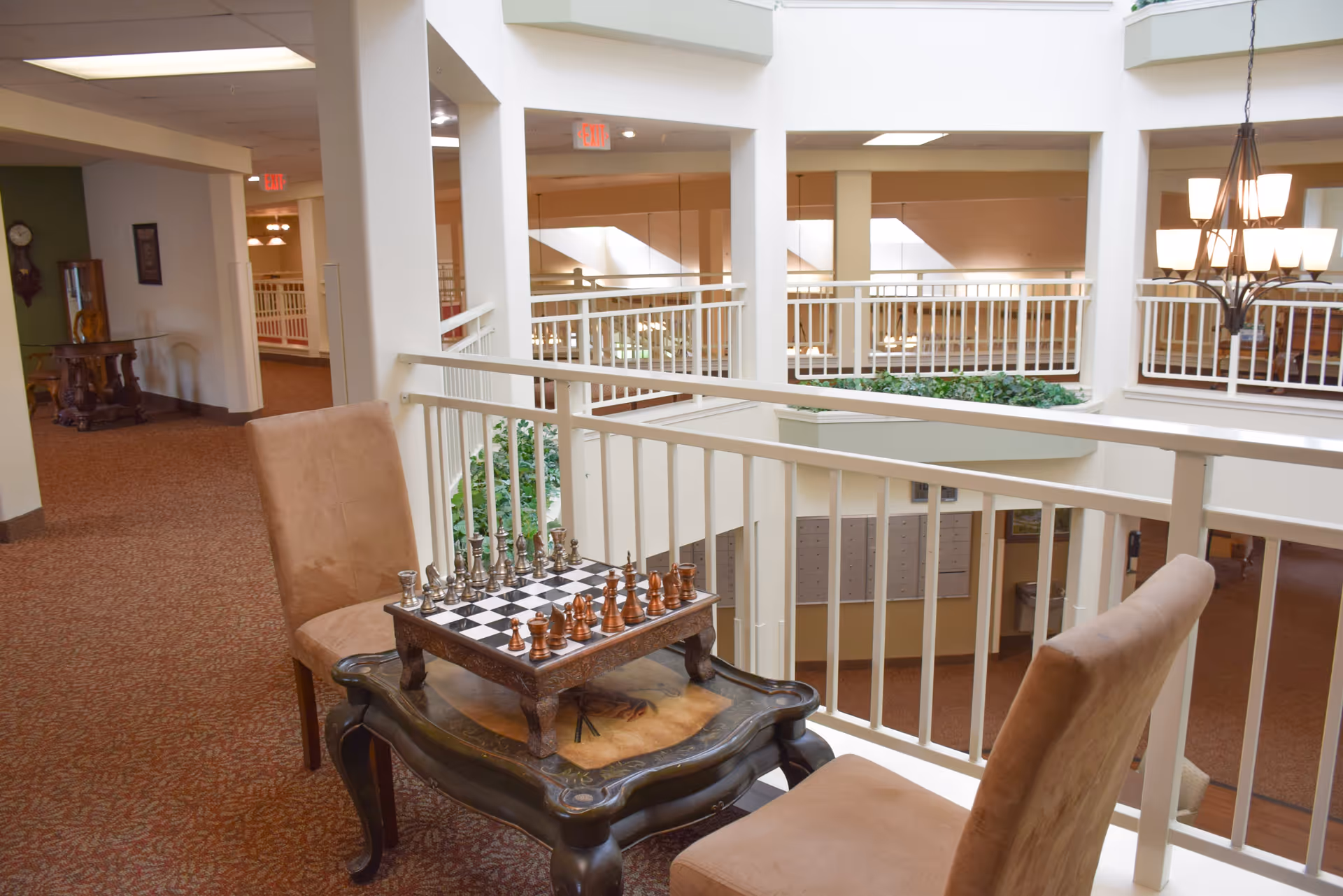 Indoor seating area in a senior living facility with two beige chairs and a small wooden table holding a chessboard with chess pieces set up. The area overlooks a lower level with white railings and a chandelier hanging from the ceiling.