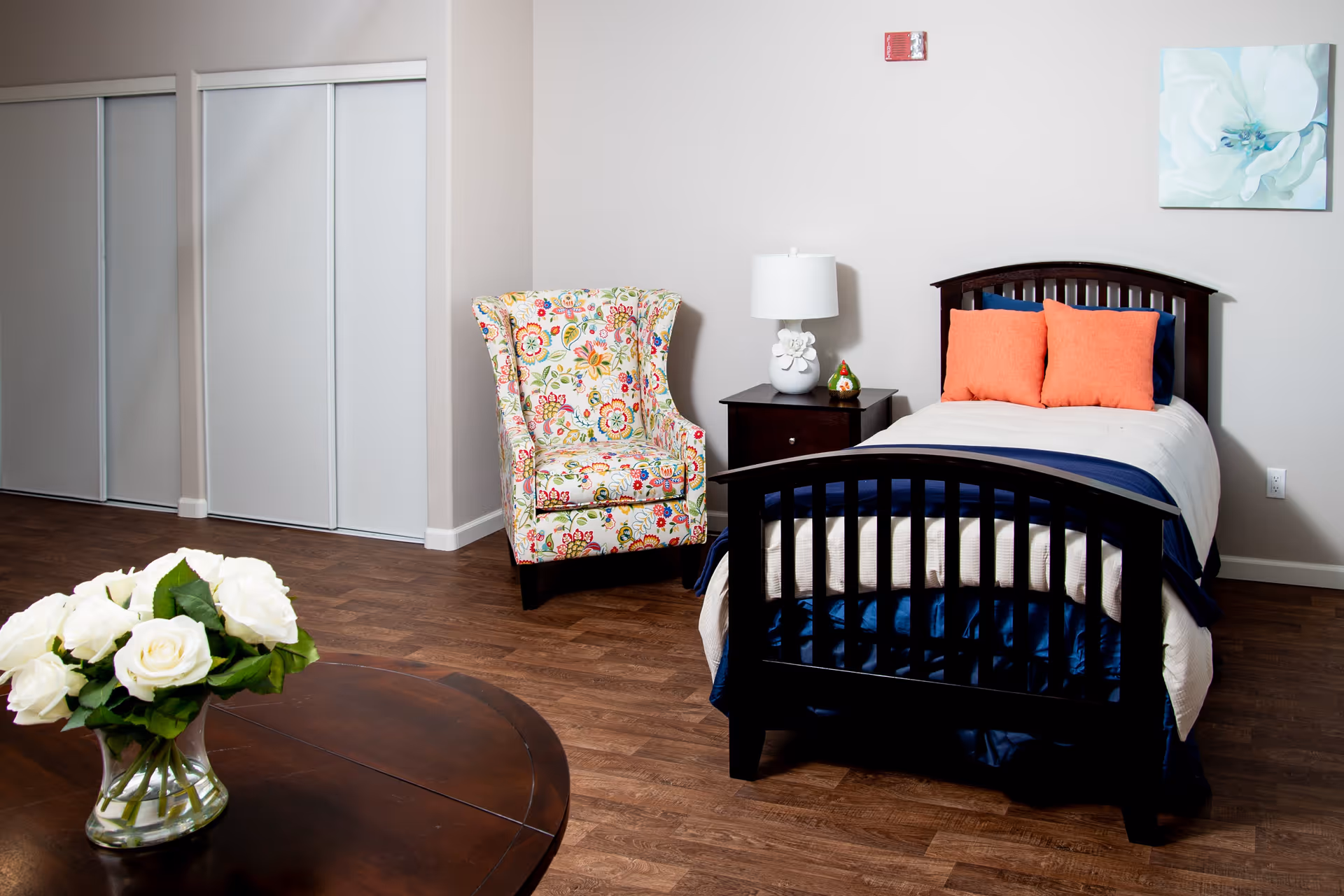 A senior living bedroom featuring a single dark wood bed with white and navy bedding and two orange pillows. Next to the bed is a dark wood nightstand with a white lamp and a small decorative item. A colorful floral armchair is positioned near a wall with white sliding closet doors. A round wooden table with a vase of white roses is in the foreground. The room has wooden flooring and light-colored walls with a floral painting hanging above the bed.
