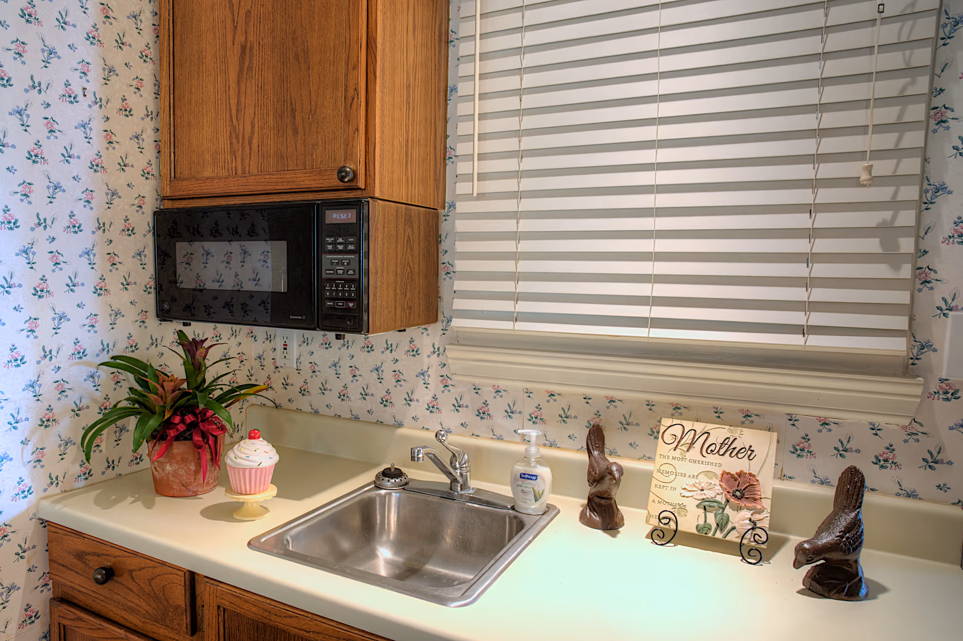 A kitchen countertop with a stainless steel sink, a soap dispenser, a potted plant with a red ribbon, a decorative cupcake, two bird figurines, and a sign that reads 'Mother The Most Cherished Memories Are Kept In A Mother's Heart'. Above the countertop is a wooden cabinet with a black microwave, and a window with white blinds. The walls have floral wallpaper.