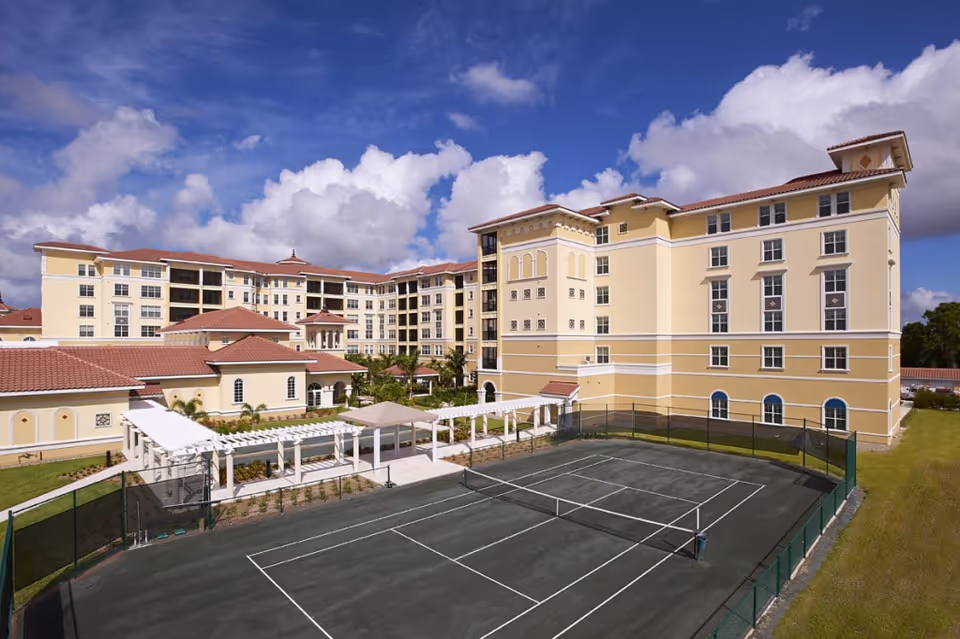 Large Mediterranean-style senior living building with a tennis court and pergola in the foreground under a blue sky.