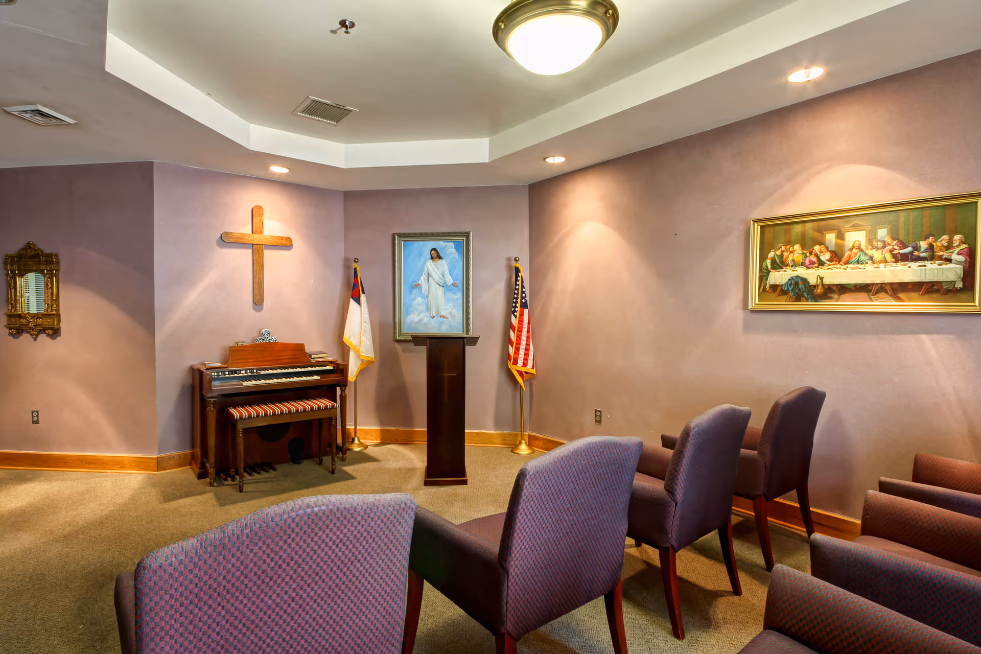 A small chapel room with purple walls and carpeted floor. There are several upholstered chairs arranged facing a wooden podium with a framed picture of Jesus above it. To the left of the podium is a wooden cross mounted on the wall above an organ with a striped cushioned bench. Two flags, one Christian and one American, stand on either side of the podium. On the right wall, there is a framed painting of the Last Supper. The room is softly lit with ceiling lights.