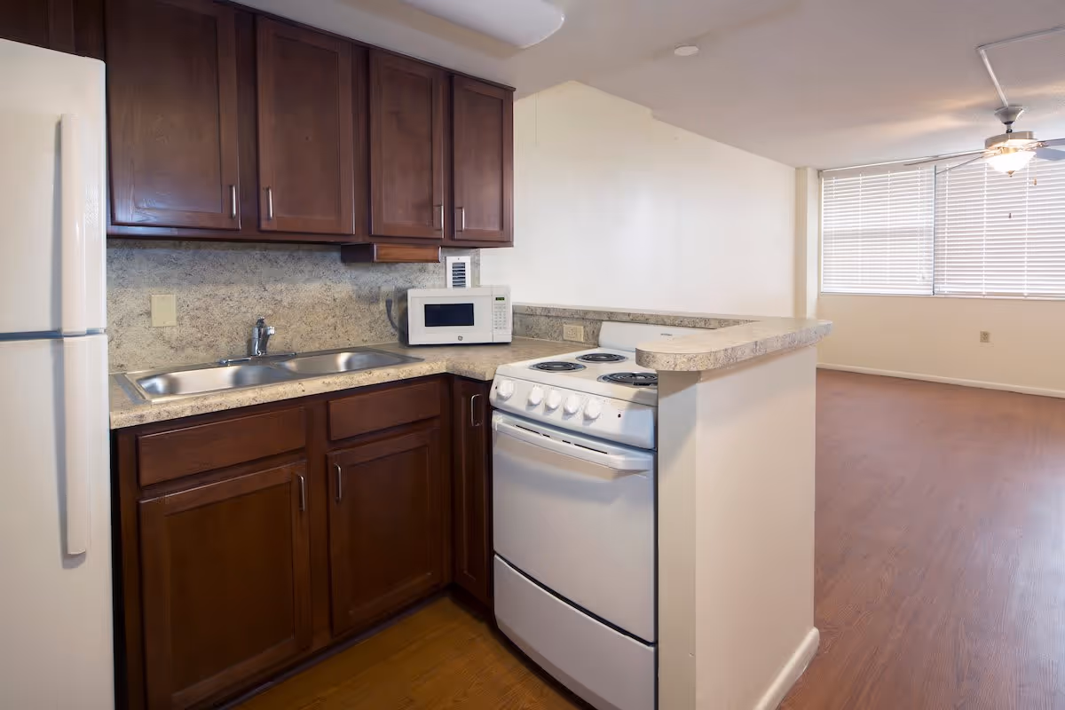 A small kitchen area with dark wooden cabinets, a white refrigerator, a white electric stove with four burners, a microwave on the countertop, and a double sink. The kitchen opens into a room with large windows covered by blinds and a ceiling fan with a light fixture.