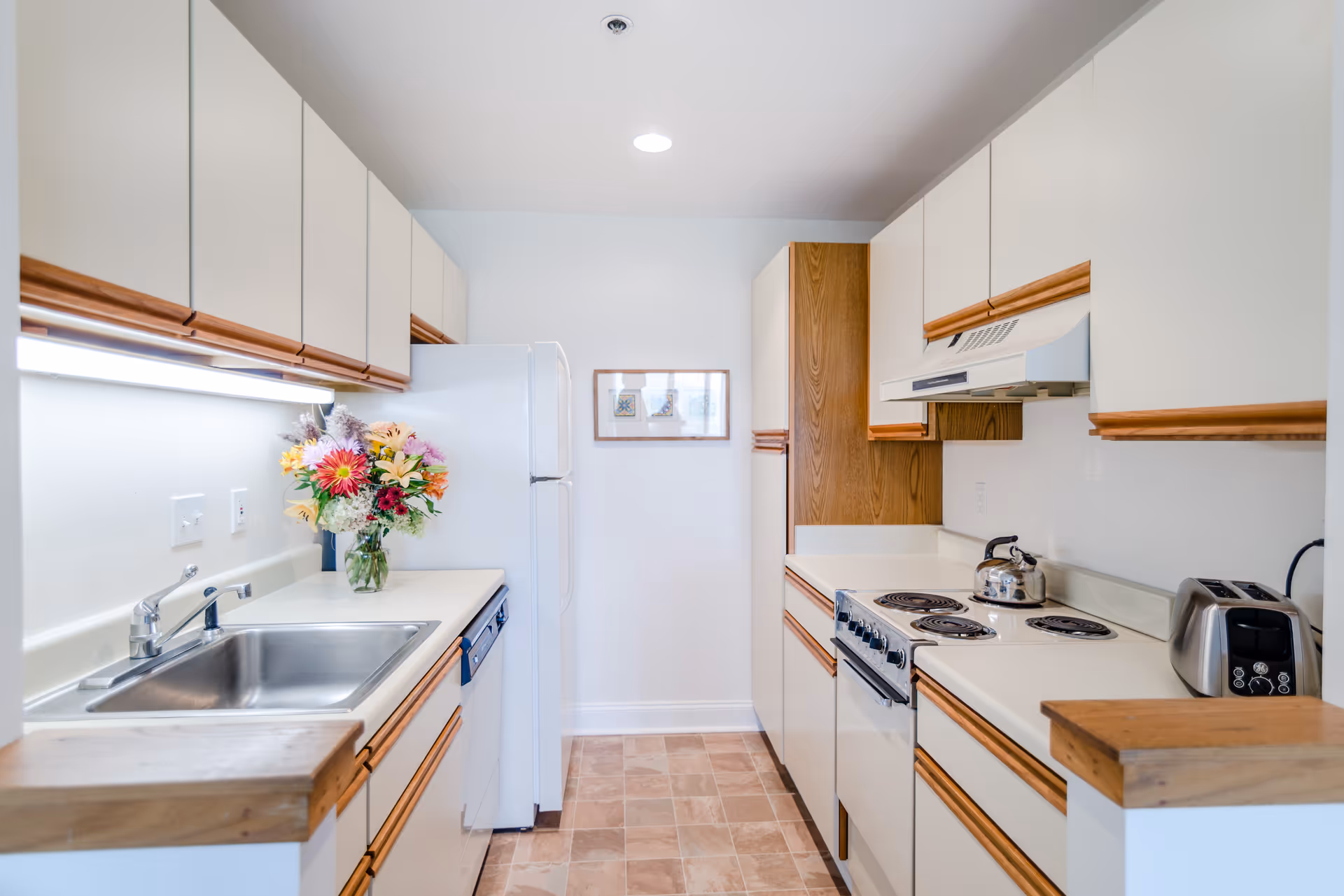 A clean, well-lit kitchen with white cabinets and wooden trim. The kitchen features a stainless steel sink, a white refrigerator, an electric stove with a kettle on it, a toaster, and a vase of colorful flowers on the counter. There is a framed picture on the far wall and tiled flooring.