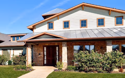 Front exterior view of a senior living facility building with stone and light-colored siding, a metal roof, multiple windows, a pathway leading to a double-door entrance, and landscaped bushes and grass in front.