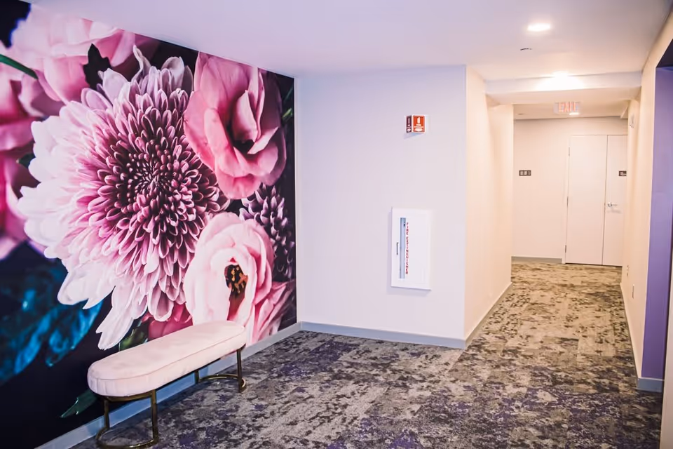 Carpeted interior hallway featuring a large pink floral wall mural, a small upholstered bench, and doors at the end.