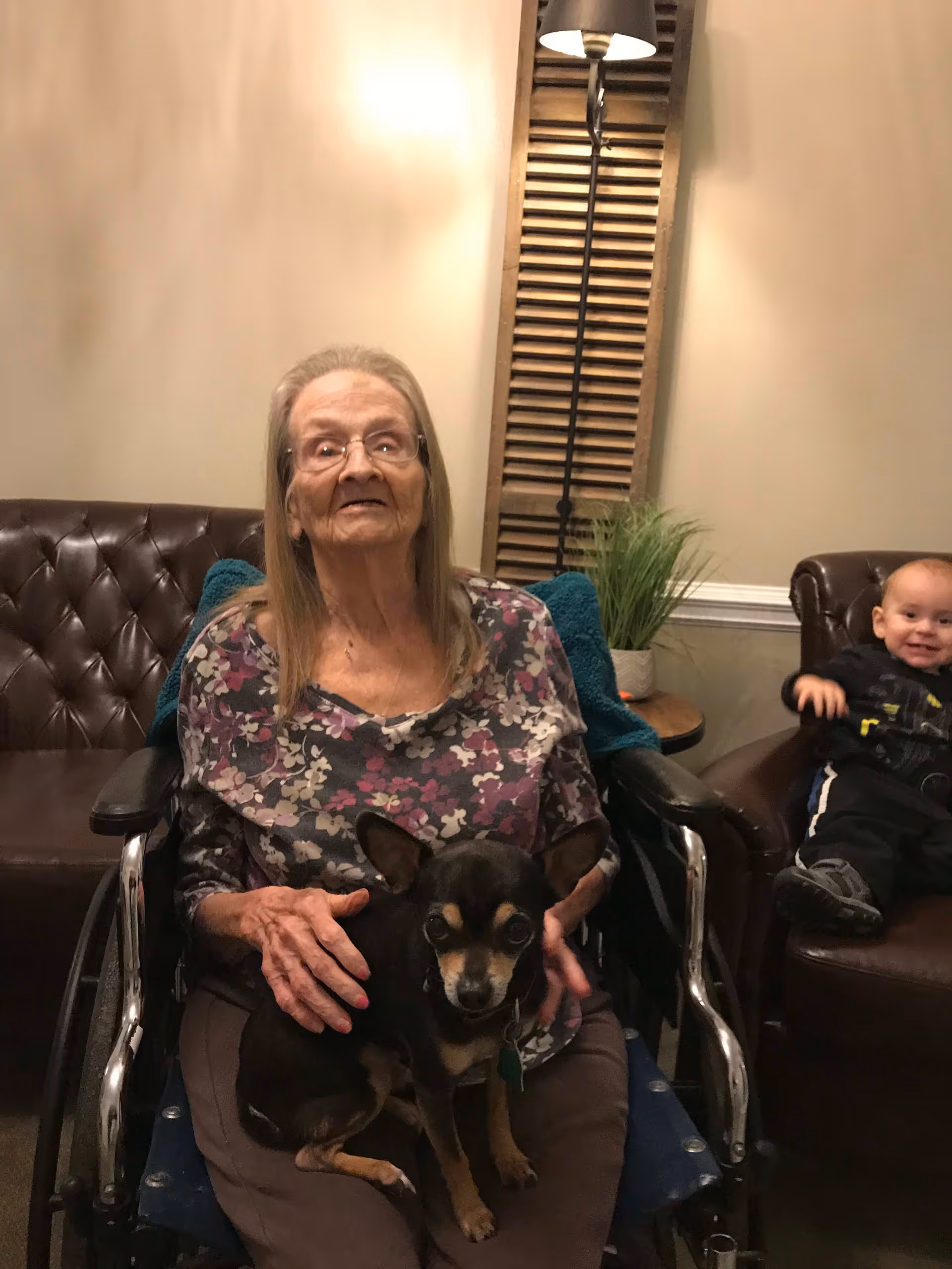 An elderly woman sitting in a wheelchair holding a small black and tan dog on her lap. Behind her is a brown leather couch with a small child sitting on it, smiling. The room has beige walls, a tall floor lamp, a wooden shutter decoration, and a small round table with a potted plant.