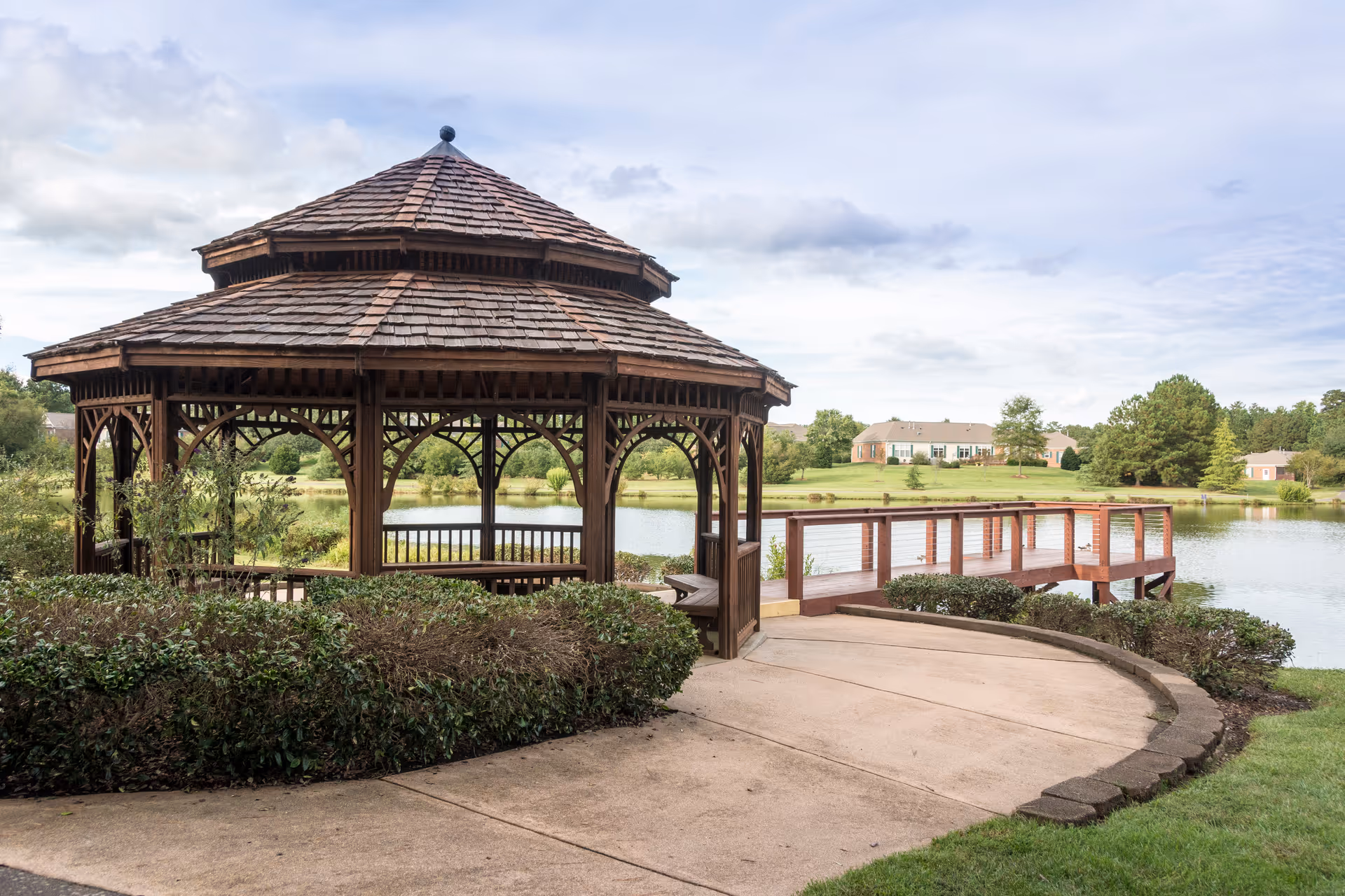 A wooden gazebo with a shingled roof situated near a pond, surrounded by greenery and bushes, with a wooden dock extending over the water and houses visible in the background under a partly cloudy sky.