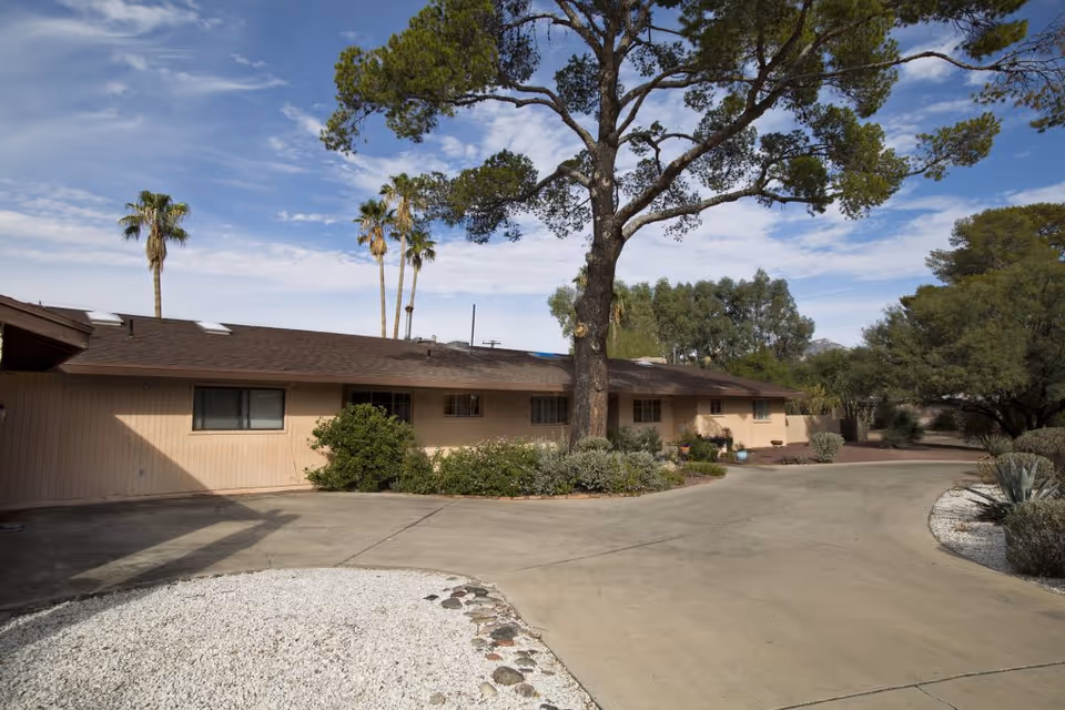 Exterior view of a single-story building with a brown roof and beige walls surrounded by trees and shrubs under a partly cloudy sky. A large tree stands in front of the building, and a curved driveway leads up to it with landscaped areas featuring white gravel and various plants.