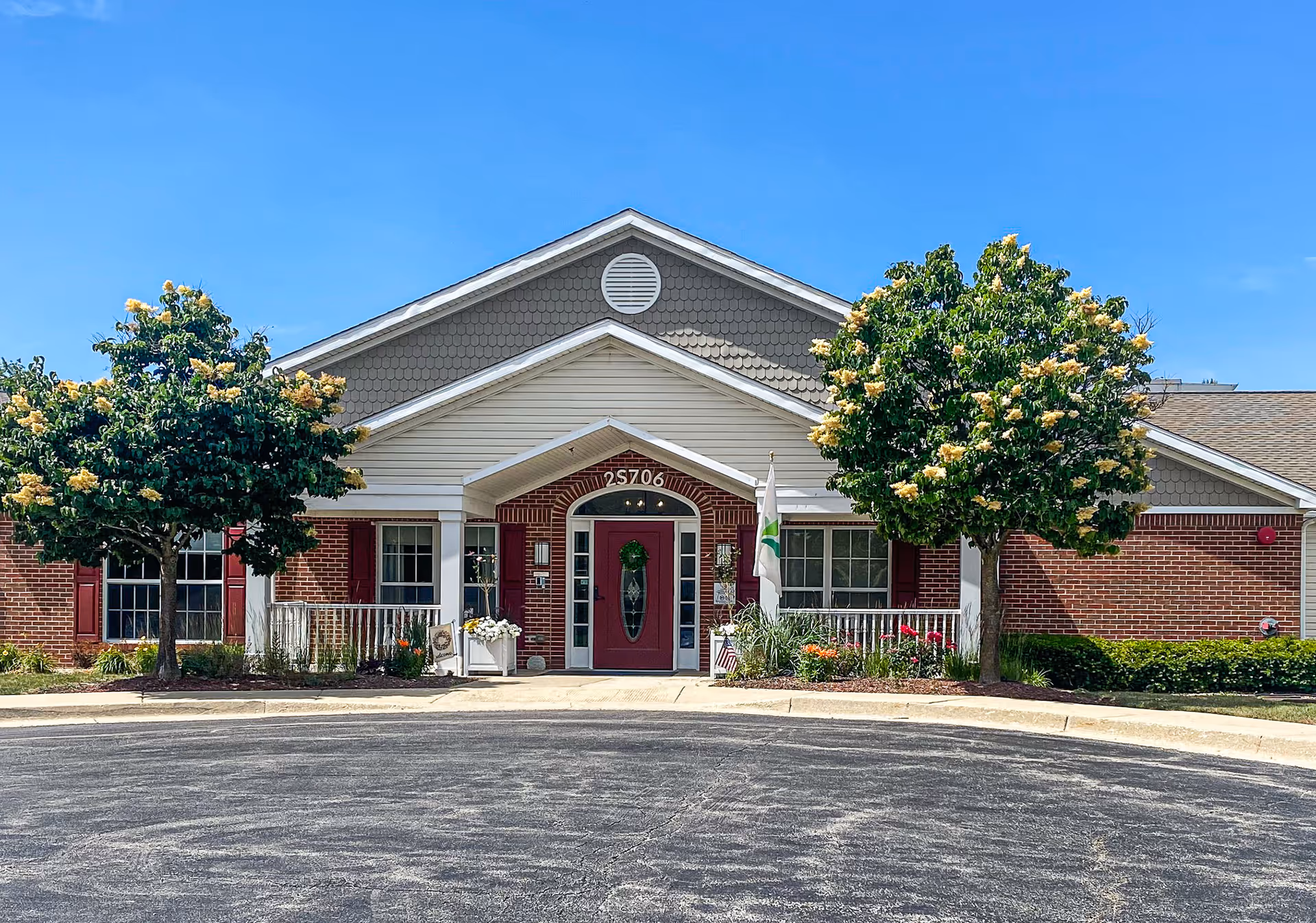 Front entrance of a single-story brick memory care building with a centered red door, two flowering trees, and a circular driveway.