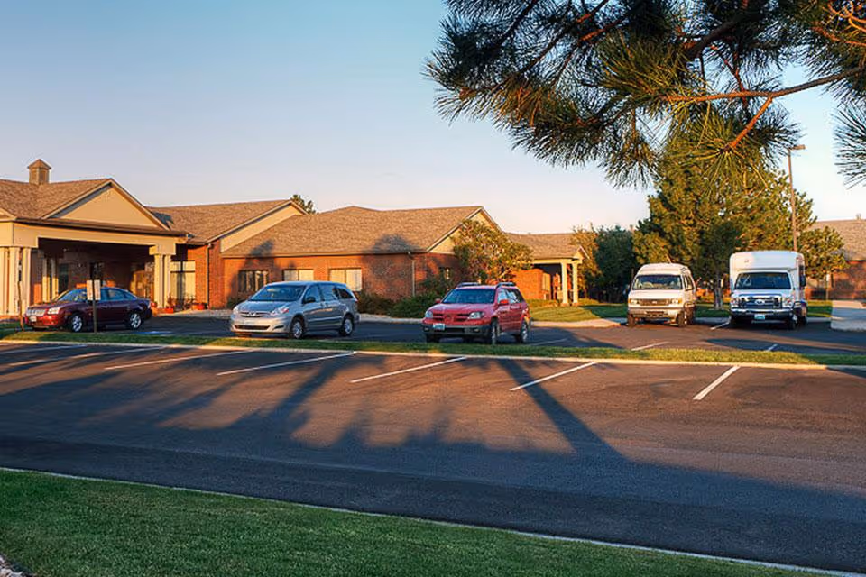 Exterior view of a single-story brick building with a covered entrance, several parked cars, and trees around the parking lot during daylight.