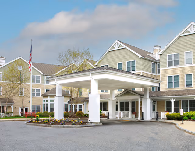 Front exterior view of a senior living facility named Mary Ann Morse at Heritage, featuring a covered entrance with white pillars, a circular driveway, landscaped flower beds, and an American flag on a flagpole. The building has multiple windows and a beige and green exterior with white trim.
