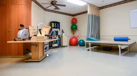 A rehabilitation room with exercise equipment including a stationary bike, therapy table with a blue bolster, various exercise balls, and hula hoops. The room has beige walls, a ceiling fan, and wooden cabinets along one wall.