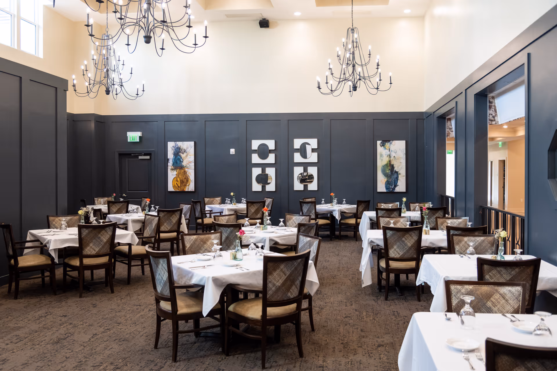 Bright, formal dining room with multiple set tables, chandeliers, and dark paneled walls.
