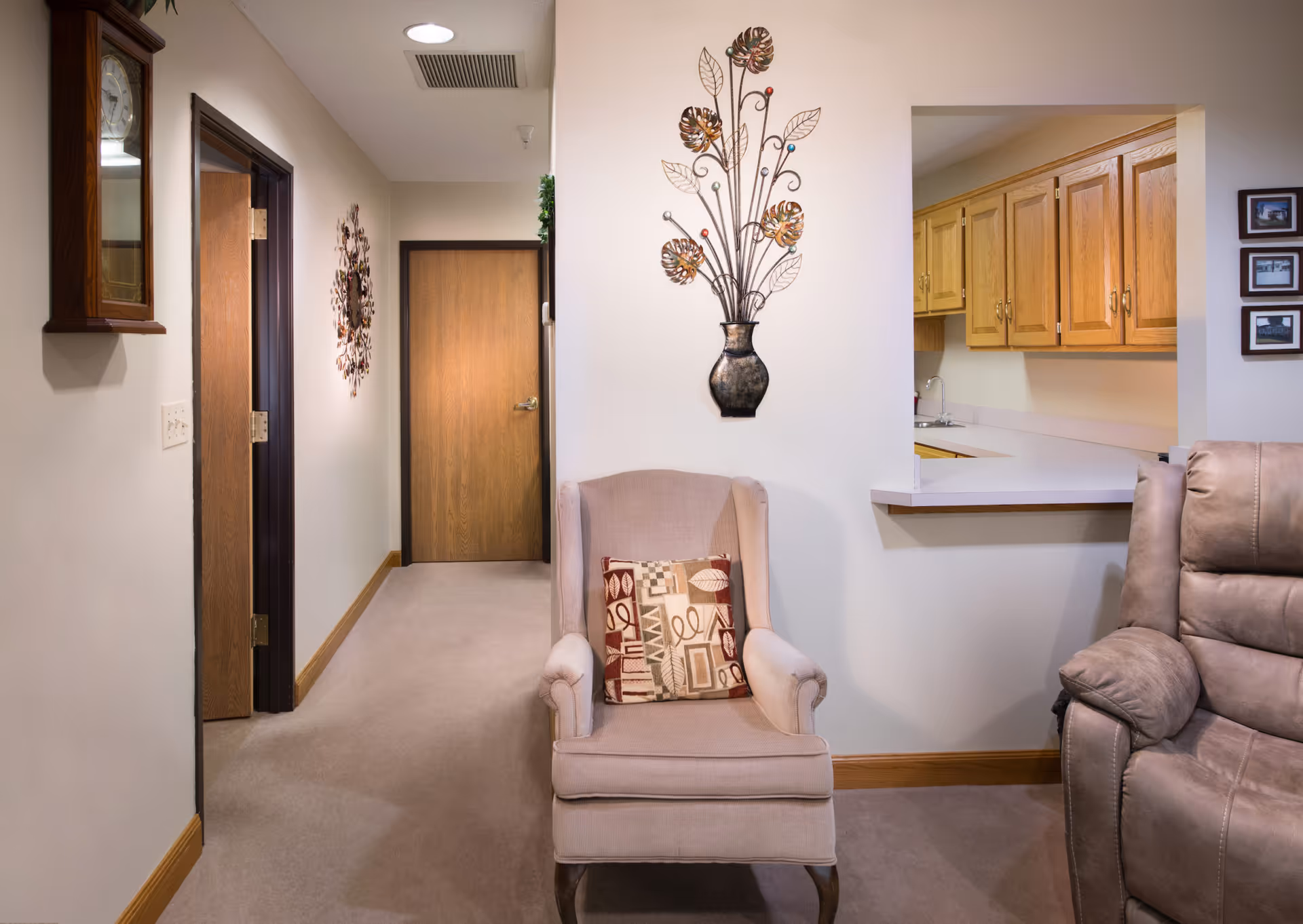 Interior view of a senior living facility showing a hallway with wooden doors, a beige armchair with a patterned cushion, a partial view of a kitchen with wooden cabinets, and a brown recliner chair. The walls are decorated with a metal wall art piece shaped like a vase with flowers and a wall clock.