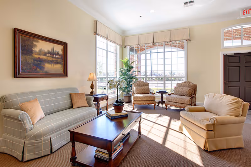 A bright and cozy living room with a plaid sofa, two patterned armchairs, and a beige armchair arranged around a wooden coffee table. Large windows with beige valances let in natural light, and there is a framed landscape painting on the wall. A small side table with a lamp and potted plants add to the inviting atmosphere.