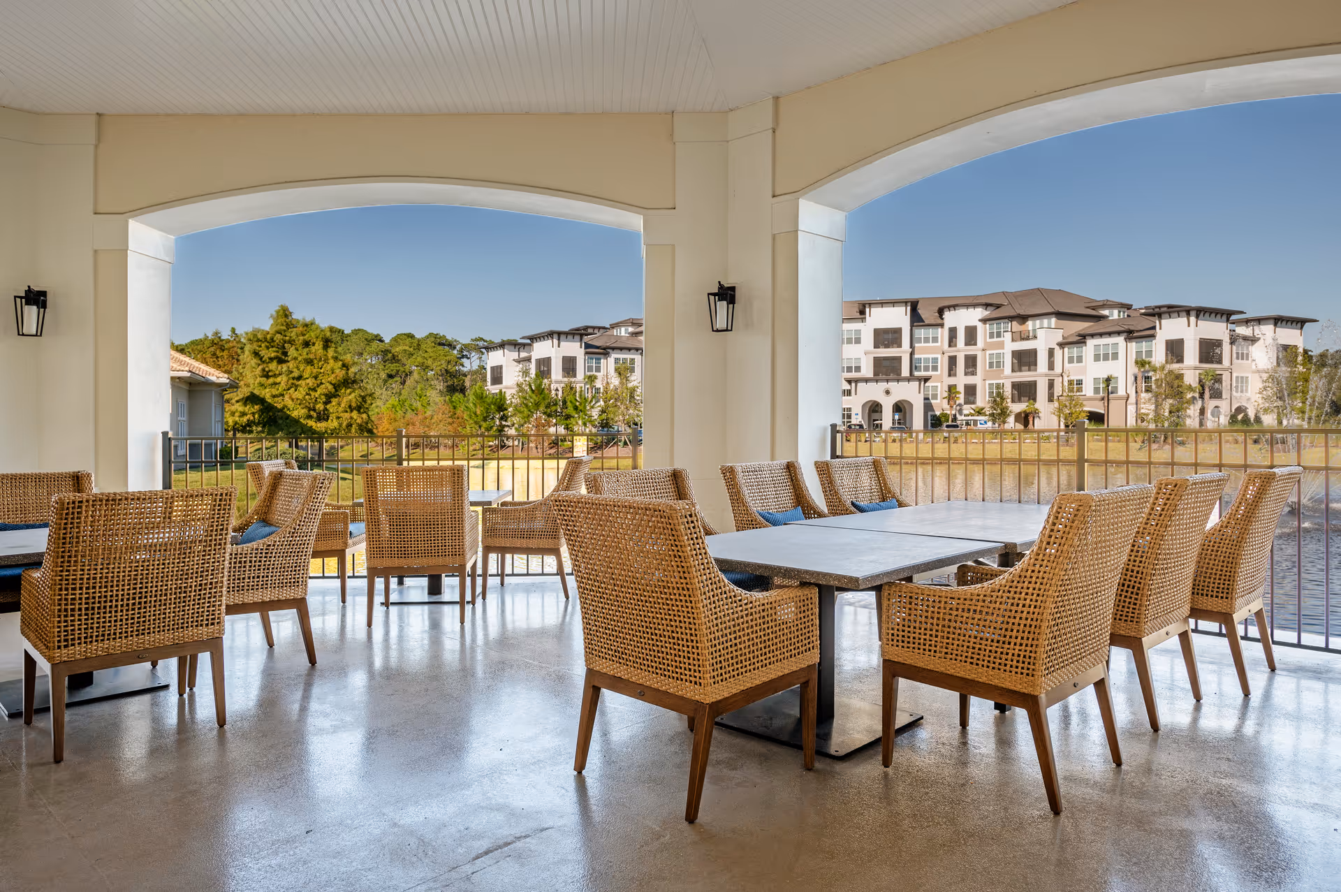 Covered outdoor seating area with multiple wicker chairs and tables overlooking a landscaped area with trees, a pond, and a multi-story residential building in the background under a clear blue sky.