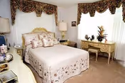 A cozy bedroom with a neatly made bed featuring a floral bedspread and multiple pillows. The room has two large windows with decorative valances, a vintage-style dresser with a matching chair, a table lamp, and various decorative items on the dresser. The carpet is beige, and the walls are light-colored.
