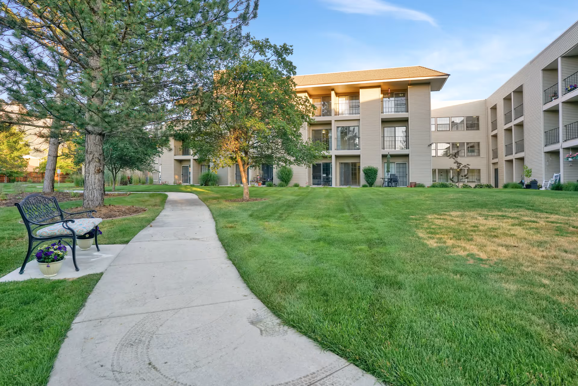 A paved walkway curves through a well-maintained grassy courtyard with trees and a bench with flower pots. Surrounding the courtyard are multi-story residential buildings with balconies and large windows under a clear blue sky.