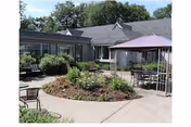 Outdoor courtyard area at a senior living facility with a circular garden bed in the center, surrounded by concrete walkways. There are several chairs and tables with umbrellas placed around the courtyard. The building with multiple windows and a gray roof surrounds the courtyard, and trees are visible in the background.