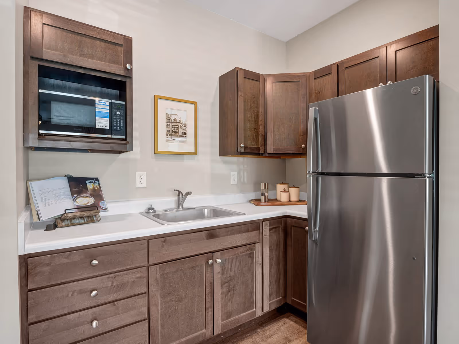 A small kitchen area with wooden cabinets, a stainless steel refrigerator, a built-in microwave, a sink, and a countertop with a cookbook and some decorative containers.