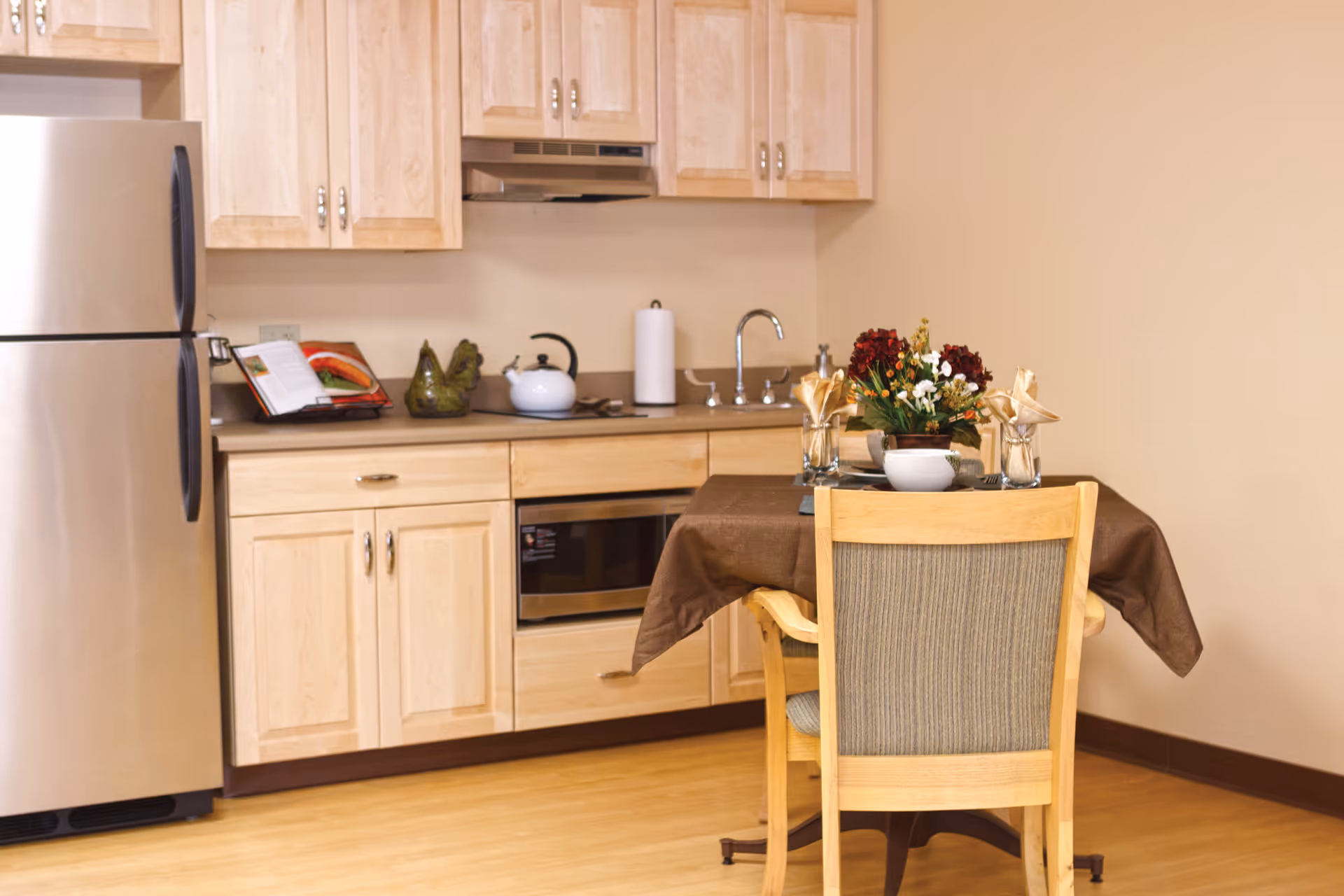 A small kitchen area with light wood cabinets, a stainless steel refrigerator, a microwave, and a sink. In front of the kitchen is a dining table covered with a brown tablecloth, set with a floral centerpiece, napkins, and utensils. The room has light-colored walls and wooden flooring.