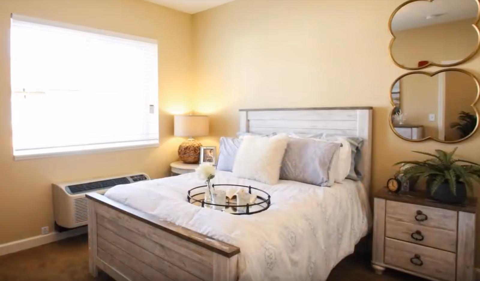 A cozy bedroom with a wooden bed frame and white bedding, decorated with multiple pillows including a fluffy white one. A round tray with a flower vase and candles is placed on the bed. There is a nightstand with a lamp and a framed photo next to the bed, and a dresser with a plant and a clock. Two decorative mirrors hang on the wall above the dresser. A window with blinds allows natural light into the room.