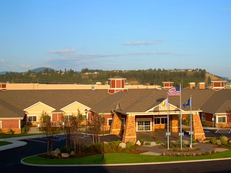 Exterior view of Life Care Center of Post Falls, a single-story building with a large entrance supported by stone pillars. Three flagpoles with flags are in front of the entrance. The building is surrounded by landscaped greenery and a paved driveway, with hills and trees visible in the background under a clear blue sky.