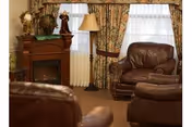 Cozy living room with brown leather armchairs, a floor lamp, patterned curtains, and a wooden cabinet with decorative items.