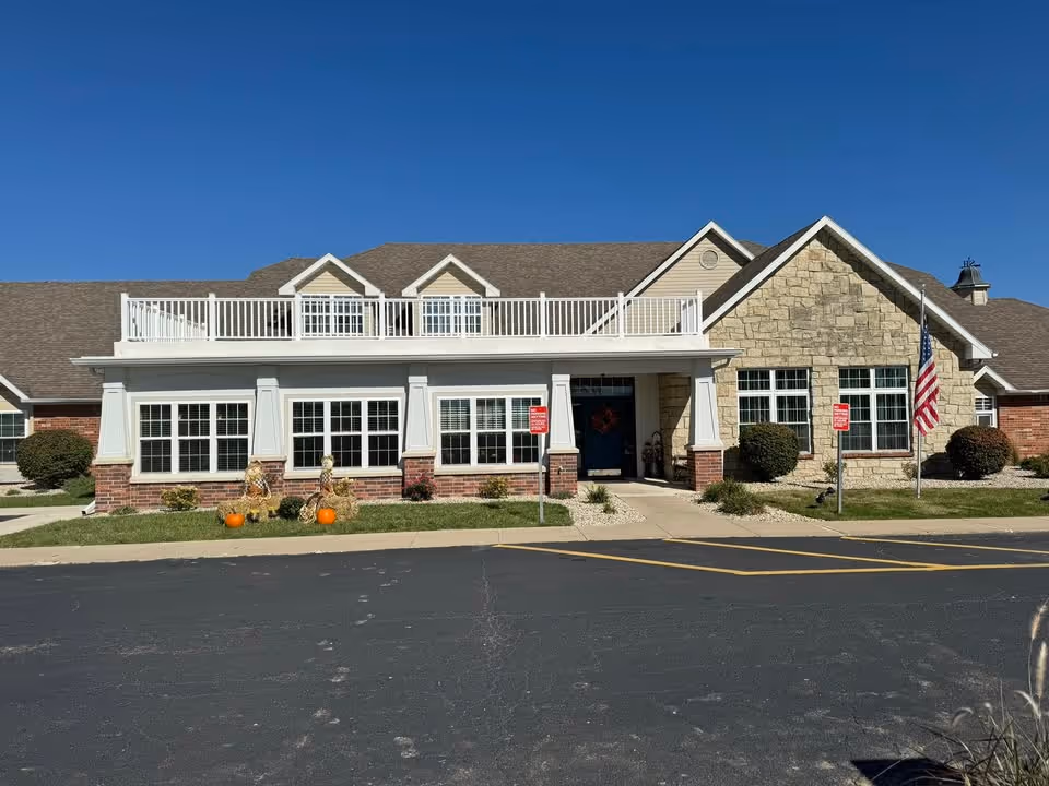 Exterior front view of a single-story senior living facility building with a combination of brick and stone facade, white railings on the roof, multiple windows, an American flag on a pole, and fall decorations including pumpkins and hay bales on the lawn.