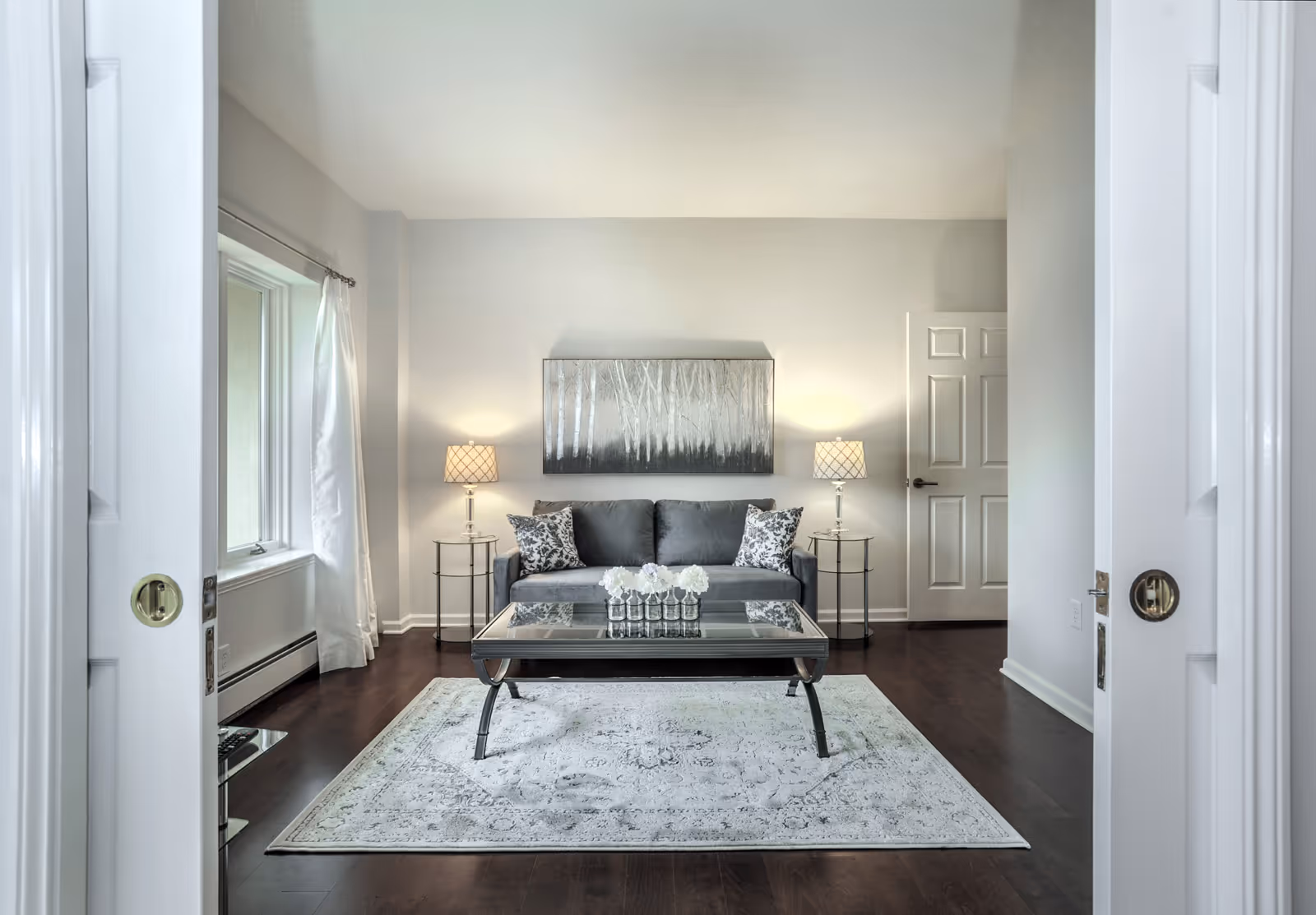 A modern living room with a gray sofa adorned with patterned pillows, two side tables each holding a lamp with a patterned shade, a glass coffee table with a decorative centerpiece, a large area rug with a subtle design, and a painting of trees on the wall behind the sofa. The room has dark hardwood floors, white walls, and a window with white curtains on the left side.