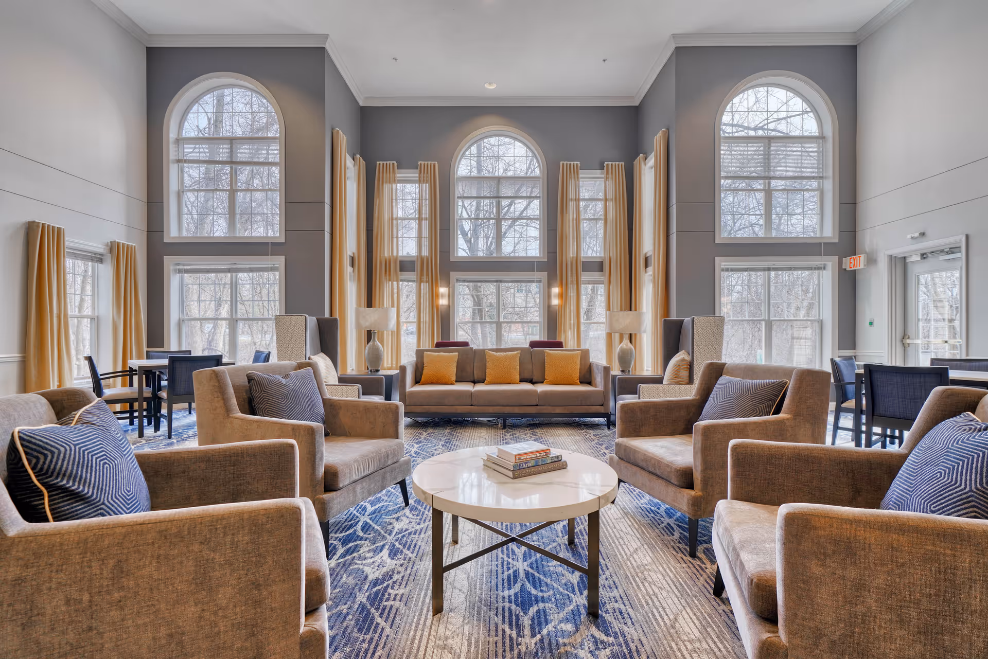 Spacious senior living lounge with tall arched windows, sofas and armchairs arranged around a central coffee table.